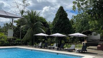Una piscina con sillas de descanso y sombrillas, rodeada de abundante vegetación y un cielo nublado.