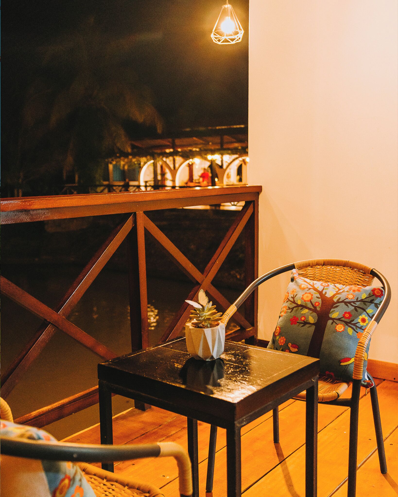 A wooden table with a small plant and a candle, a wicker chair with a floral cushion, and a hanging light fixture.