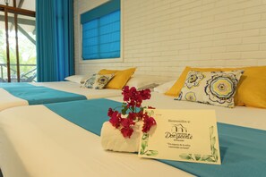 A hotel room with two beds, blue and yellow bedding, a welcome card, and a decorative plant.