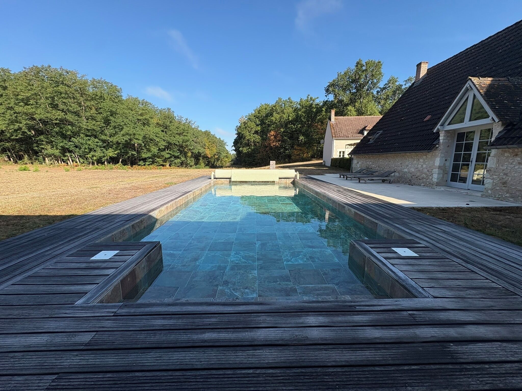 A rectangular swimming pool with a wooden deck surrounding it, set against a backdrop of trees and a building with a sloped roof.