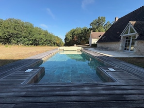 A rectangular swimming pool with a wooden deck surrounding it, set against a backdrop of trees and a building with a sloped roof.