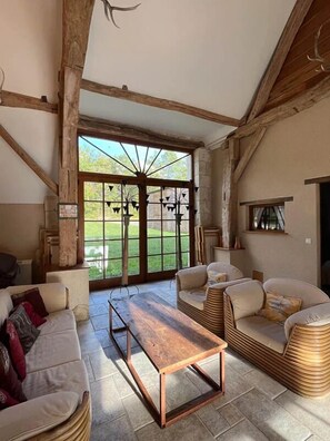 A rustic interior with a wooden ceiling, a large window, a wooden table, and two wicker sofas.