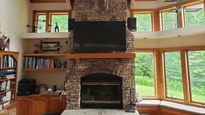 A stone fireplace with a mounted deer head, a television above it, and large windows with a view of greenery. - ML03-Lakefront Retreat, Hot Tub, Pets Ok! New A/C! (Davis)