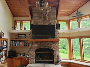 A stone fireplace with a mounted deer head, a television above it, and large windows with a view of greenery.