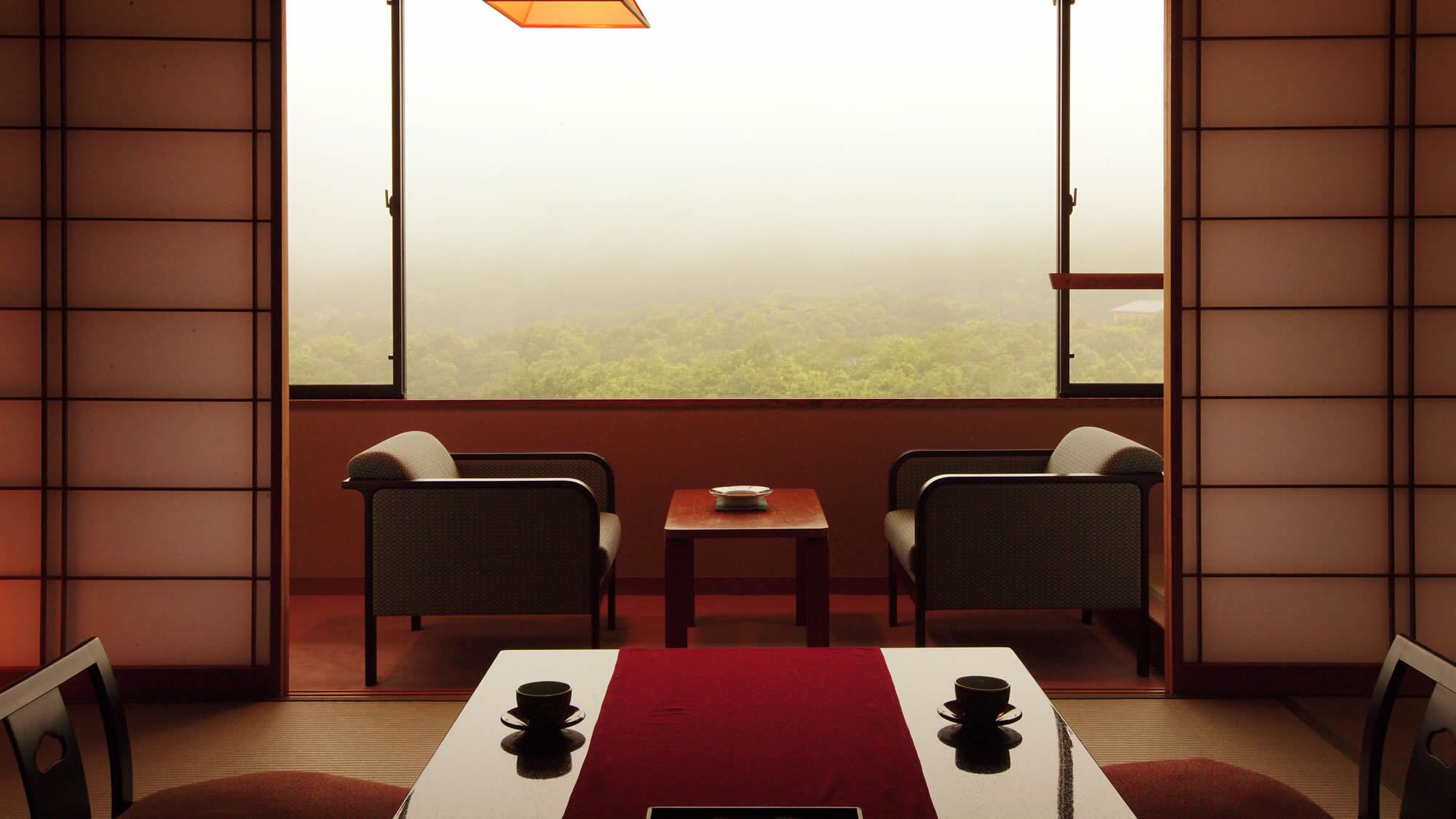 A room with tatami mat flooring, a low table with a red runner, two chairs, and a view of a forest through large windows.