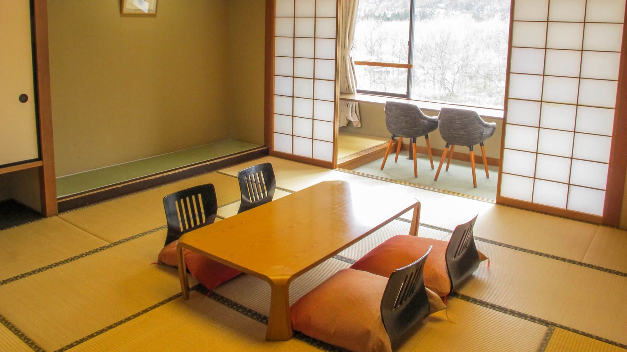 A tatami mat room with a low table and chairs, a sliding door leading to a balcony with chairs, and a view of a snowy landscape.