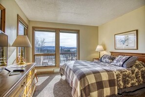 Une chambre avec une grande fenêtre, un lit recouvert d’une couverture à carreaux, une commode et un tableau encadré accroché au mur.