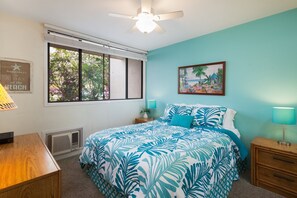 A bedroom with a bed, a desk, a chair, a ceiling fan, a window with blinds, a wall-mounted picture, and a nightstand with a lamp.