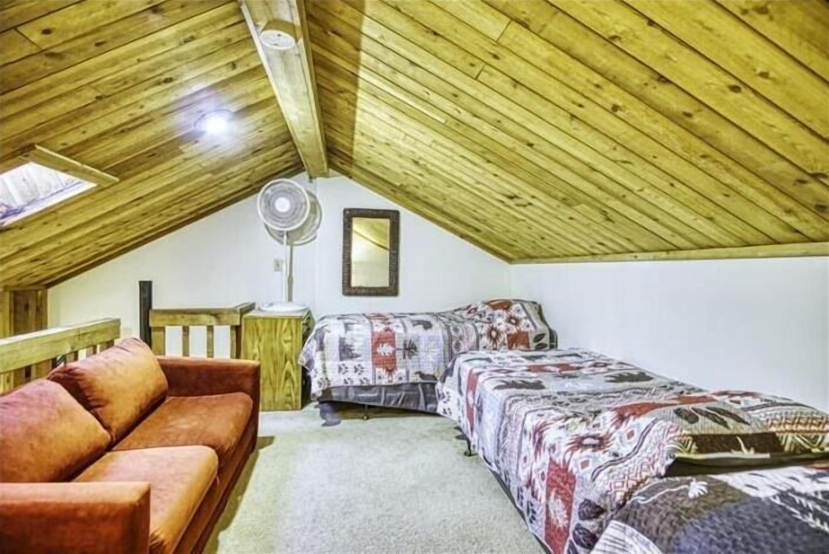 A cozy attic bedroom with a wooden ceiling, a single bed with patterned bedding, a red sofa, and a small table with a fan.