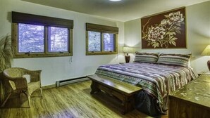 Ein Schlafzimmer mit einem großen Bett, zwei Fenstern mit Jalousien, einem Sessel, einem Nachttisch und einer Wandlampe.