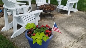 Eine Terrassenfläche mit weißen Adirondack-Stühlen, einer Feuerstelle und einem Topfpflanzenbeet mit einer amerikanischen Flagge.