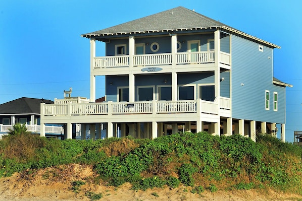 A multi-story beachfront house with balconies, a flat roof, and a sandy foreground.