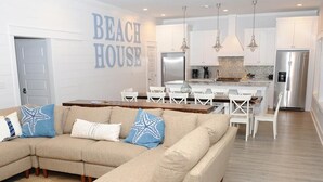 A modern living room with a beige sectional sofa, a dining area with white chairs, a kitchen with white cabinets, and a stainless steel refrigerator. - Amazing Grace: 7  BR, 5  BA House in Port Bolivar, Sleeps 25 (Port Bolivar)