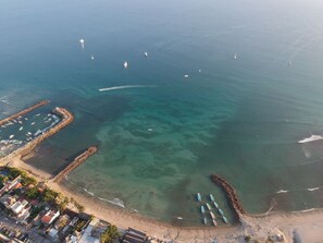Vue aérienne d’une zone côtière avec des eaux cristallines, un port de plaisance et une plage bordée de bâtiments et de palmiers.