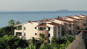Un hotel costero con varias edificaciones, cada una con techo de tejas rojas y paredes blancas. El hotel está rodeado de vegetación exuberante y palmeras, con vista al mar y una isla distante.