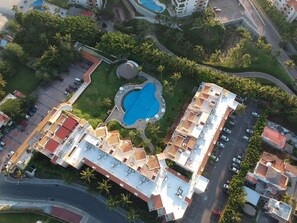 Vue aérienne d’un hôtel composé de plusieurs bâtiments, avec une piscine centrale et de nombreux espaces verts environnants.