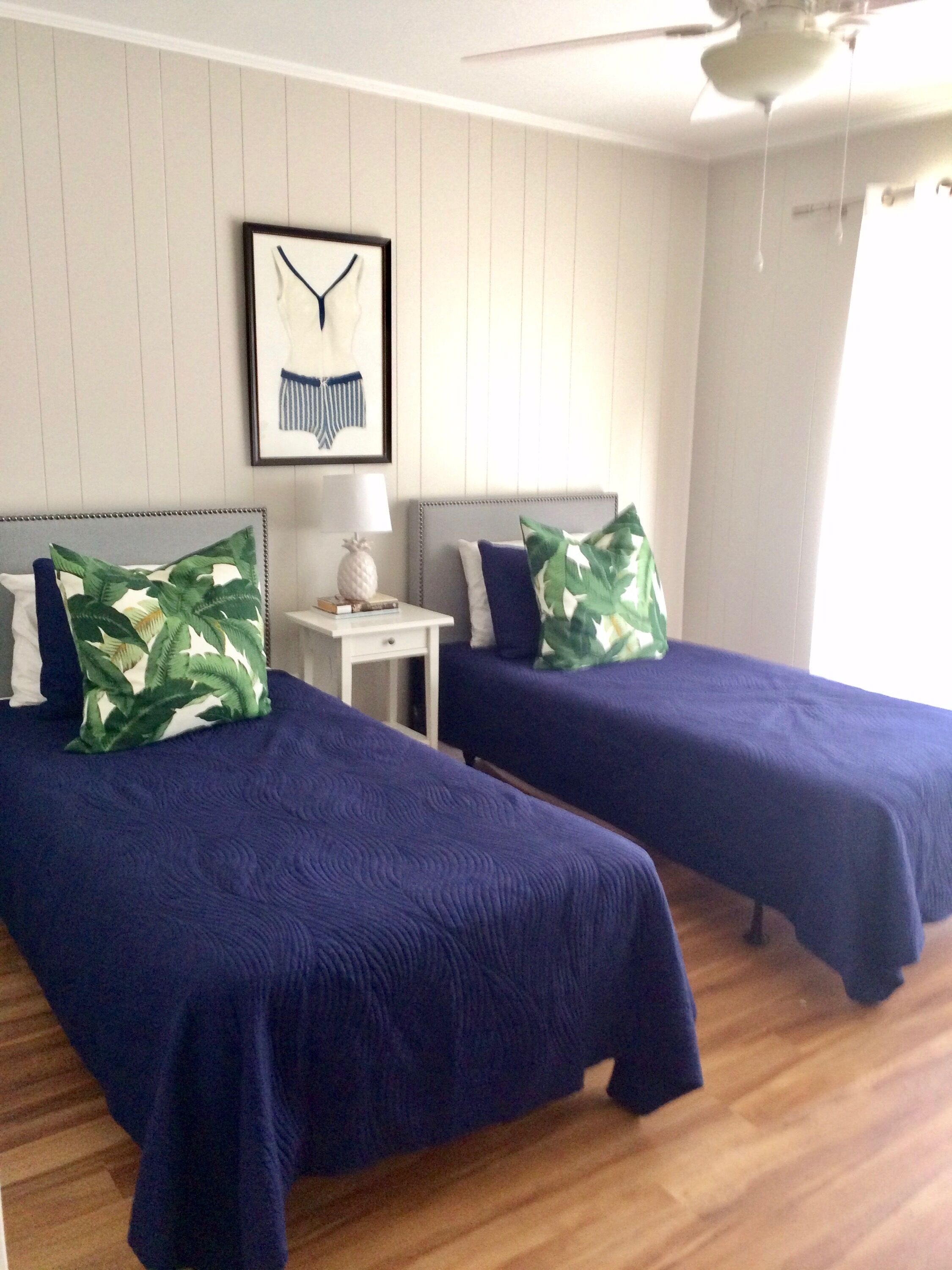 Una habitación con dos camas, cada una con una colcha azul y un cojín estampado con hojas verdes. Hay una mesita de noche con una lámpara y un cuadro en la pared.
