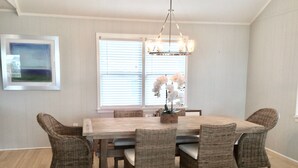 A dining area with a large wooden table surrounded by wicker chairs, a chandelier, and a window with blinds.