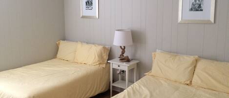 A room with two single beds, each with a white bedspread and matching pillows. There is a small bedside table with a lamp between the beds.