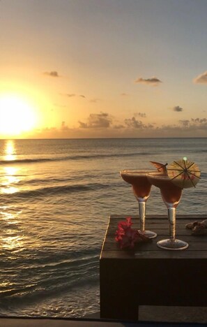 Une vue sur le coucher de soleil, avec deux boissons posées sur une table en bois, au bord de l’océan.