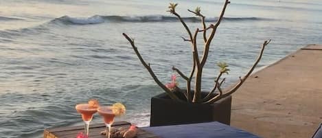 A beachside lounge with two cushioned chairs and a small table, a potted plant, and a bottle of champagne.