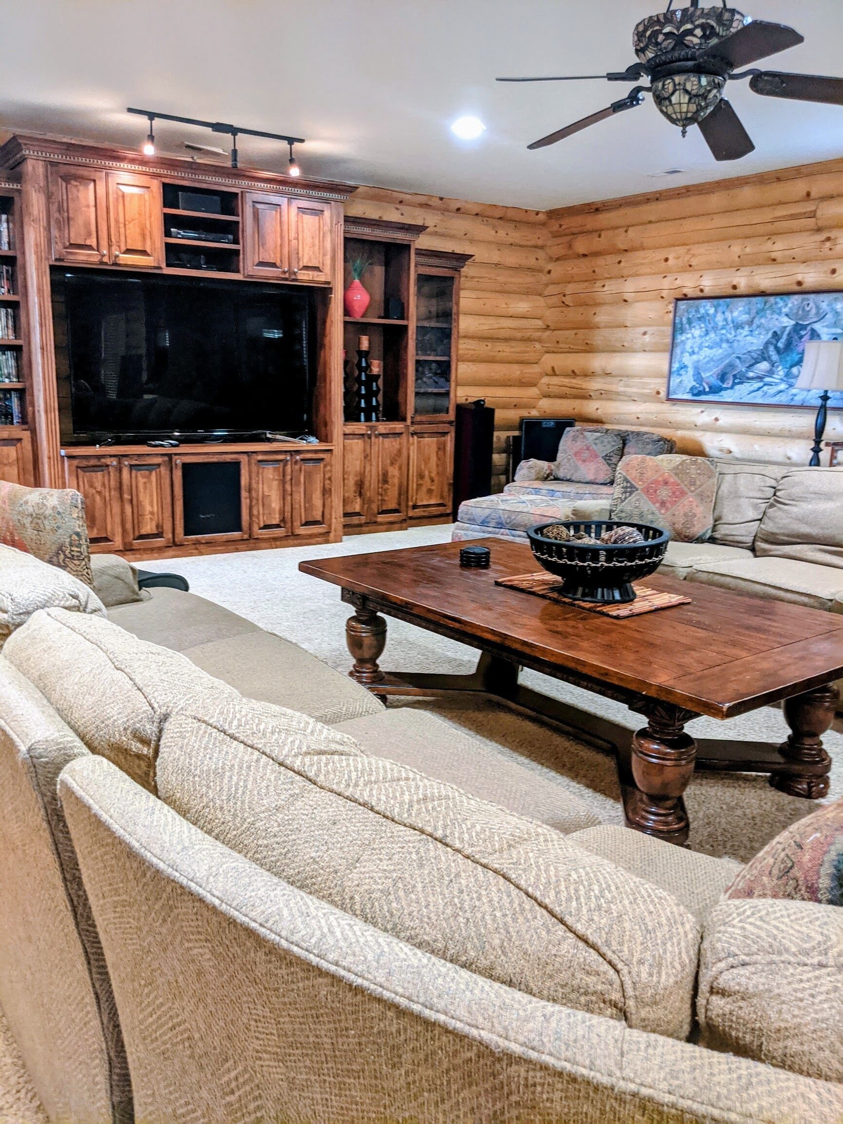 A log cabin interior with a large TV, wooden cabinets, a sofa, and a ceiling fan.