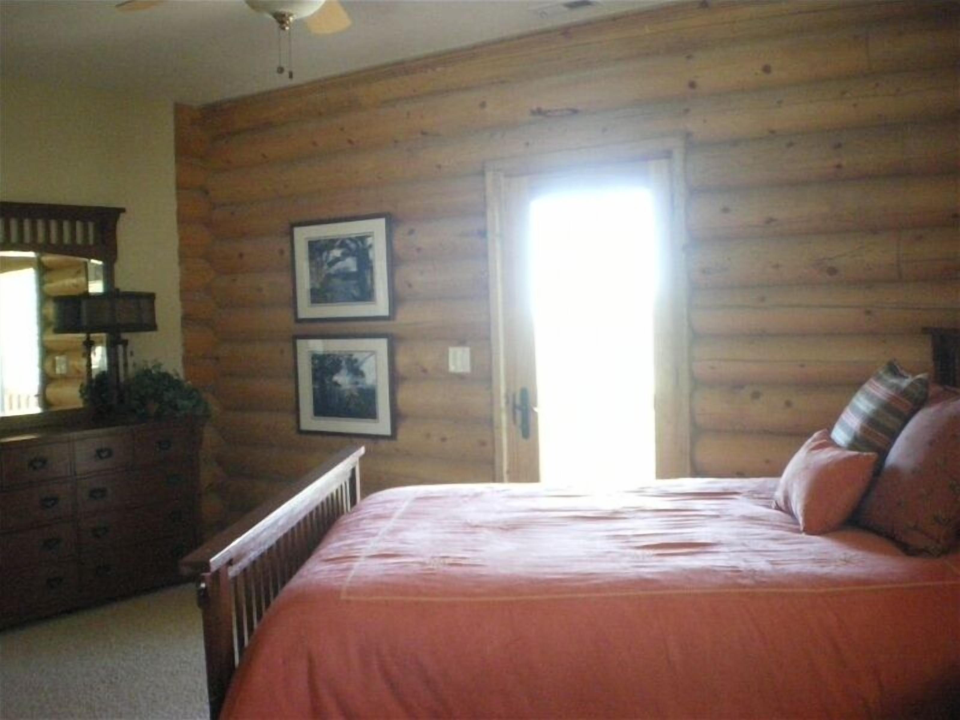 A bedroom with wooden walls, a bed with a red bedspread, two framed pictures on the wall, and a dresser with drawers.