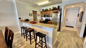 A modern kitchen with a central island, bar stools, wooden cabinets, stainless steel appliances, and a wooden floor. - Ocean Reef Beach-Front, Luxury, Condo with Awesome Balcony, Highly Rated! (Panama City Beach)