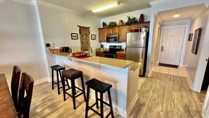 A modern kitchen with a central island, bar stools, wooden cabinets, stainless steel appliances, and a wooden floor.
