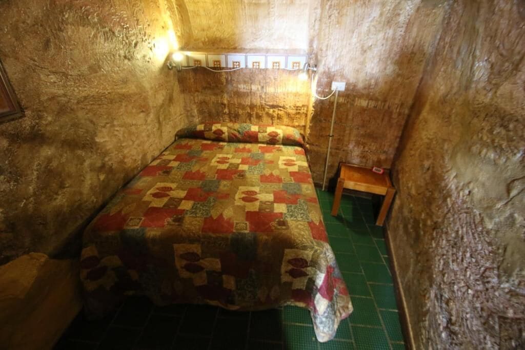 A small, rustic room with a single bed, a nightstand, and a wall-mounted light fixture.
