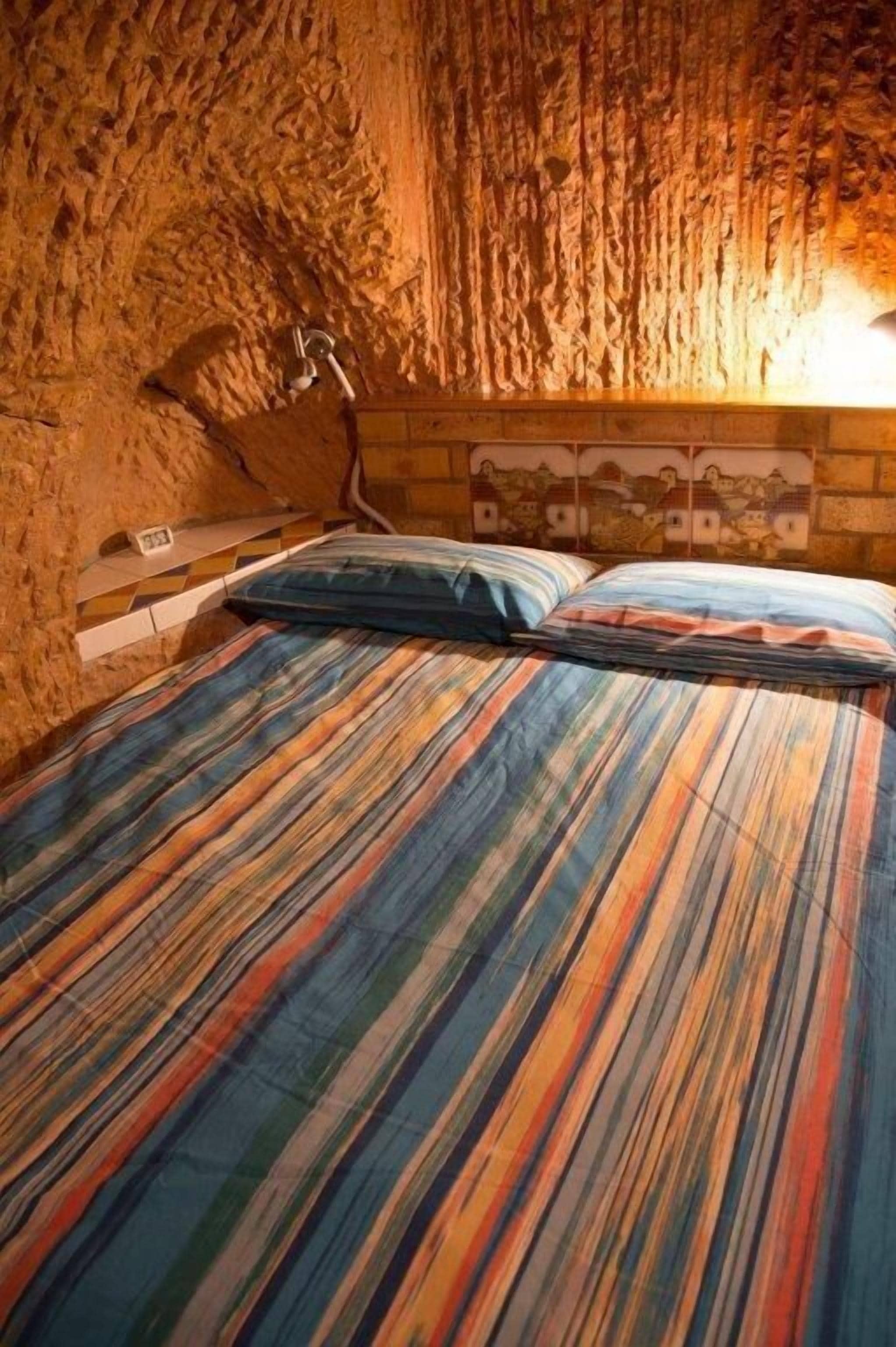 A bed with a colorful striped bedspread in a room with a textured stone wall and a wooden headboard with a cityscape mural.