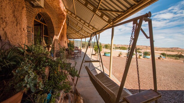 A covered outdoor seating area with rustic stone walls, a swing set, and potted plants. - The Underground Motel (Coober Pedy)