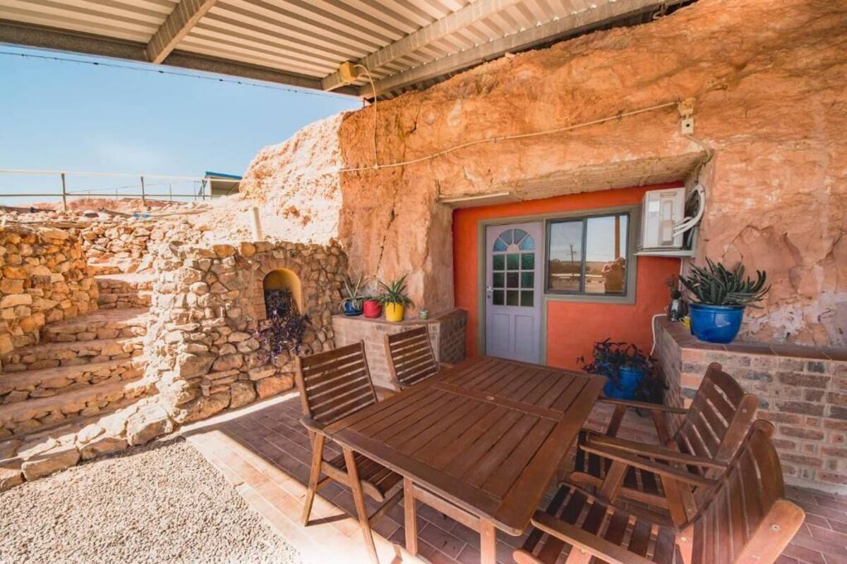 Un patio rústico al aire libre con pared de piedra, mesa y sillas de madera, y plantas en macetas.