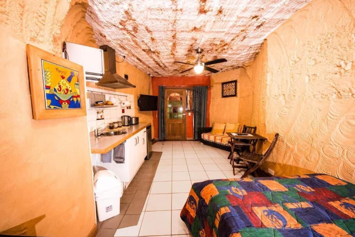 Una habitación compacta con kitchenette, una cama con colcha colorida, un ventilador de techo y una pequeña mesa.