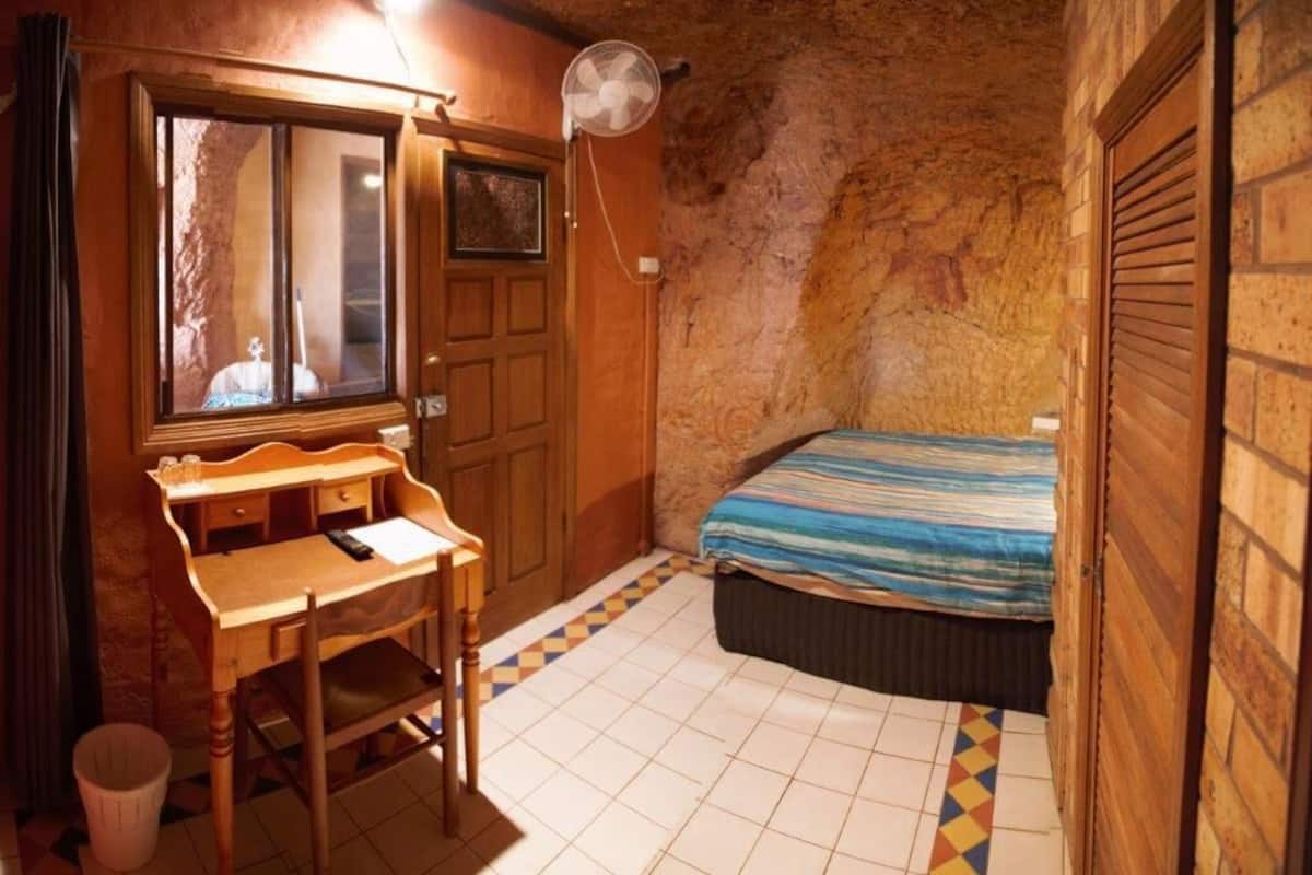 Una habitación pequeña y acogedora con un escritorio de madera, una cama con colcha a rayas, un ventilador y suelo de baldosas.