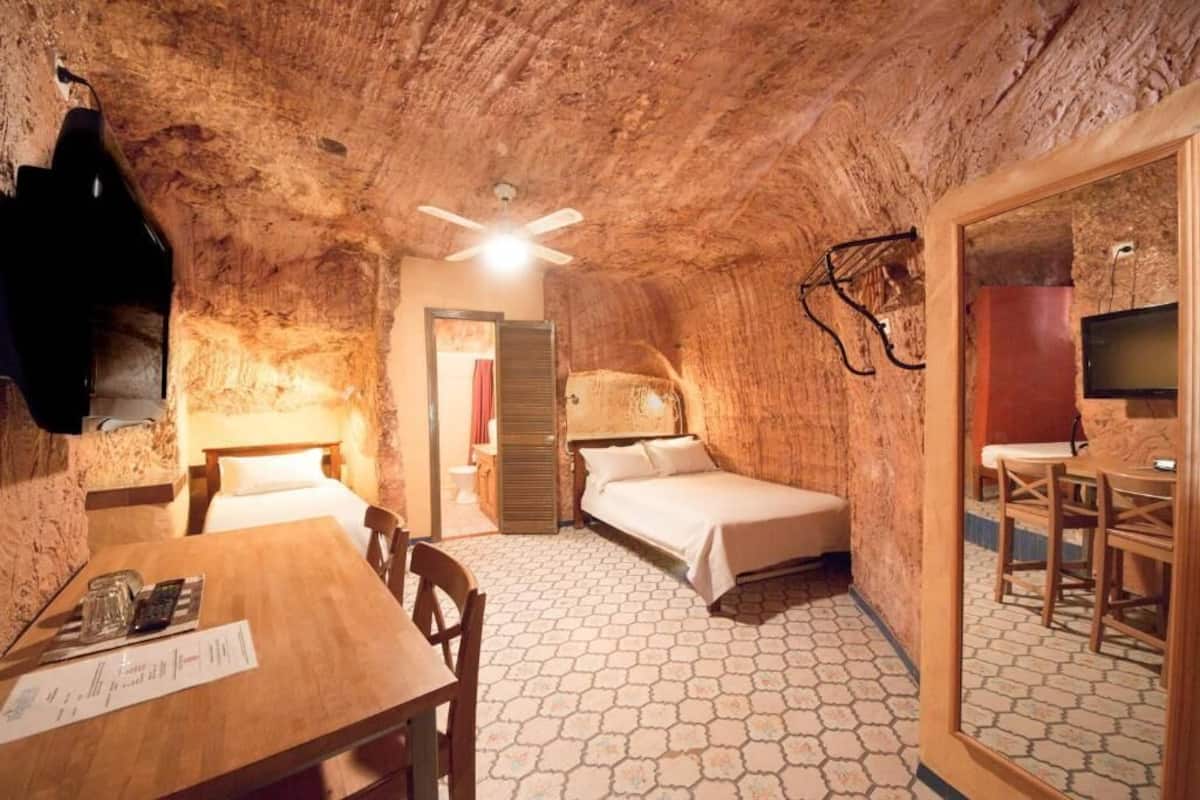 Una habitación rústica con una mesa de madera, dos camas, un ventilador de techo y un televisor.