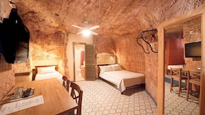 A rustic room with a wooden table, two beds, a ceiling fan, and a television. - The Underground Motel (Coober Pedy)