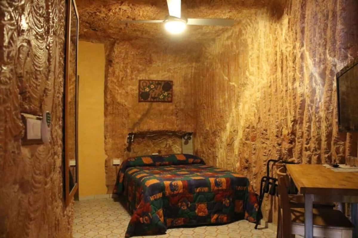 Una habitación con pared de piedra texturizada, una cama con colcha colorida, un escritorio de madera y un ventilador de techo.