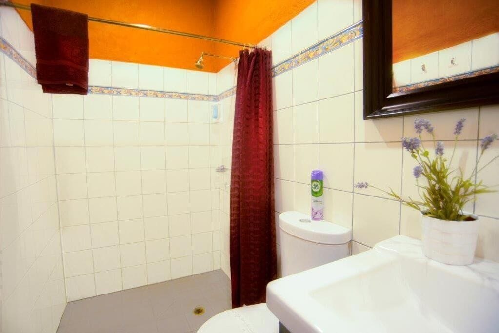 Une salle de bain avec des murs recouverts de carreaux blancs, une douche avec un rideau rouge, un lavabo blanc et un miroir encadré au-dessus du lavabo.
