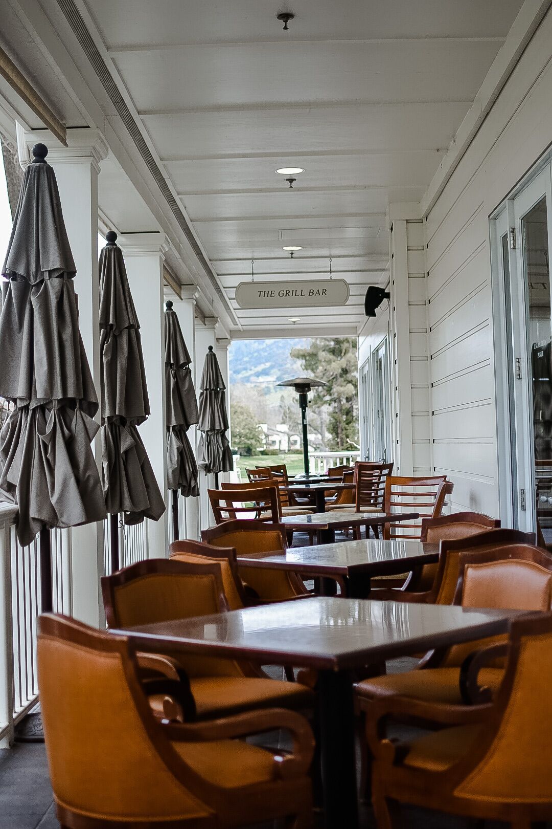 A covered outdoor dining area with tables and chairs, a sign reading "The Grill Bar", and a view of the outdoors.