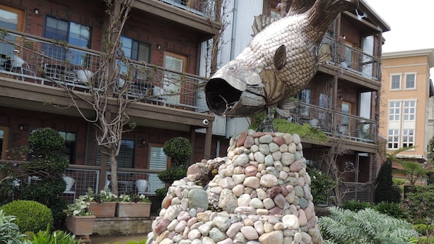 Une sculpture de poisson dans un jardin agrémenté d’une structure en pierre, entouré de bâtiments.