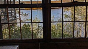 A room with a large window overlooking a lake, a wooden deck, and a view of trees and a clear sky. - Private beautiful setting, Lyme's best kept secret, Swim, Boat, Fish, Hike (Old Lyme)
