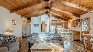 A spacious living area with wooden beams, a sofa set, a coffee table, and a kitchen in the background. - HIDDEN VISTA: Distance views from private oversized deck nestled in cool pines. (Munds Park)