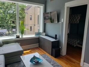 A modern living room with a sofa, a coffee table, a TV on the wall, and a window with a view of a brick building and trees. - Fulton market / Noble Square FREE parking, Laundry, 2.5 miles to Mag Mile! (Chicago)