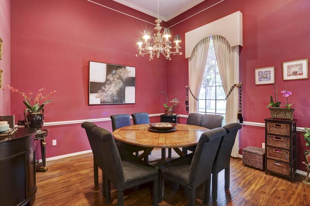 A dining room with a round wooden table, six chairs, a chandelier, and a large window with curtains.