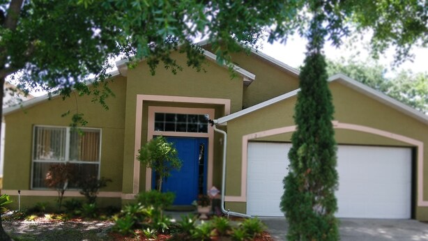 Ein eingeschossiges Haus mit blauer Haustür, einer Garage und einem kleinen Vorgarten mit Pflanzen und einem Baum.