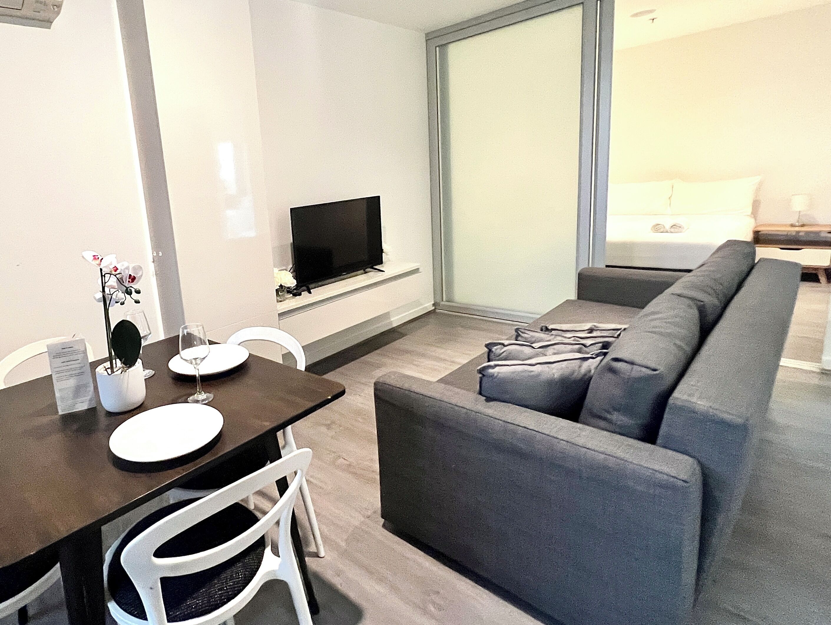 Een moderne woonkamer met een bank, een eettafel met stoelen, een flatscreen televisie aan de muur en een groot raam met uitzicht op de omgeving.