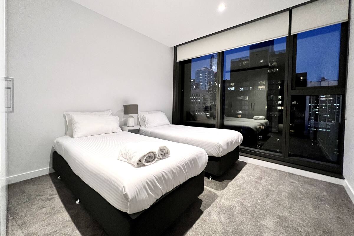 A modern hotel room with two beds, a large window with a city view, and a carpeted floor.