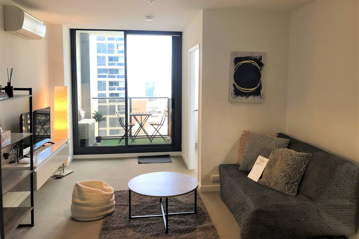 A modern living room with a grey sofa, a round coffee table, and a balcony with a view of buildings.