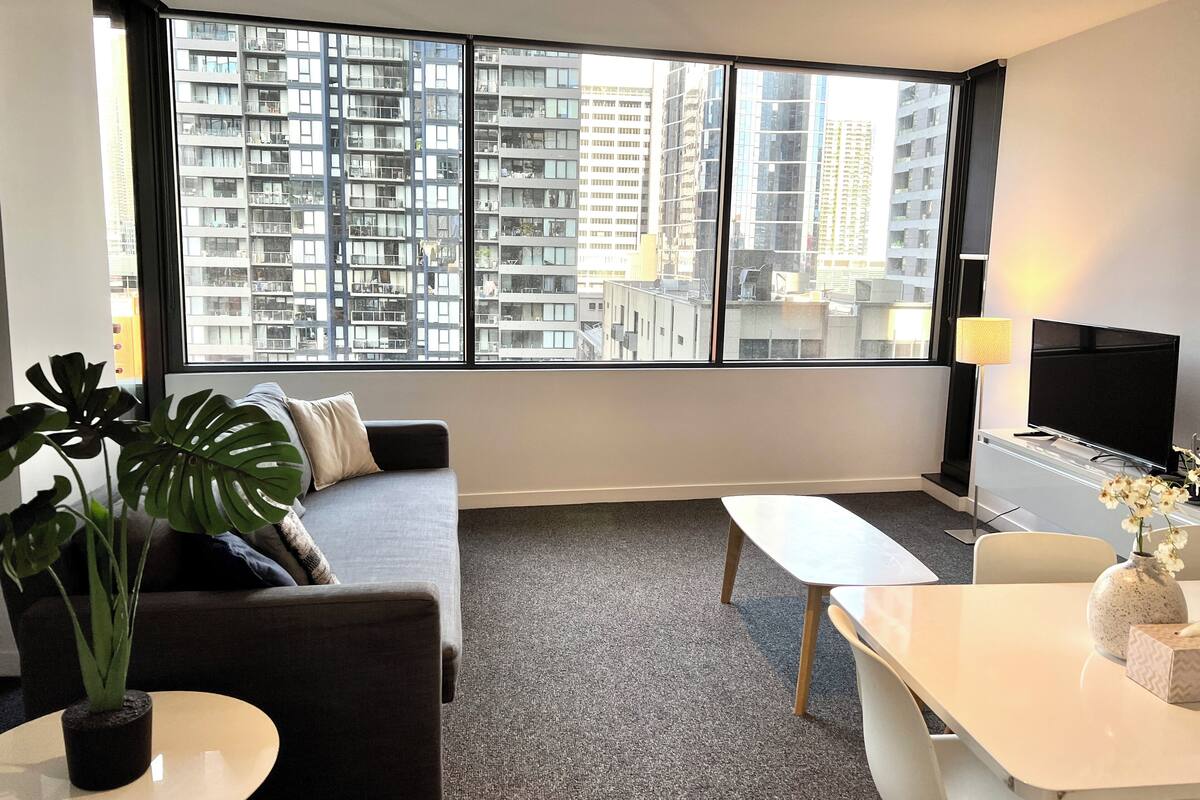 A modern living room with a sofa, a coffee table, a TV, and a large window offering a city view.